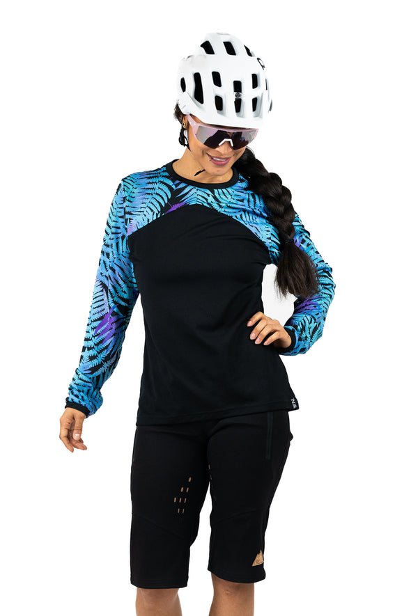 Schwarzes und blau-lila Langarm-Mountainbike-Jersey für Frauen mit floralem Farn-Print. Dargestellt an einem Modell mit Mountainbik Short, Helm und Brille.